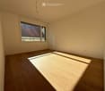 Empty room with wooden flooring and large windows allowing natural light to enter. The walls are painted a light beige color, and there are no visible furnishings or decorations.