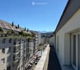 Balcony overlooking a city street with parked cars, buildings, and distant mountains under a clear blue sky.