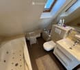 Modern bathroom with a bathtub, toilet, sink, mirror, and various toiletries. Features skylight windows and a small shelf with cups and cleaning supplies.