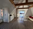 Modern attic living space with wooden beams, exposed ceiling, large windows offering a scenic view, and contemporary furniture including a sofa, dining table, chairs, and a cabinet.