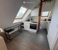 A modern, compact kitchen area with white cabinets, a built-in oven, microwave, and a small black table with a glass top. The space features wooden flooring and grey tiled flooring in the kitchen area, with skylight windows providing natural light.