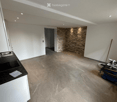 Empty room with white walls, beige tiled floor, and a small black countertop with a sink. A stone accent wall and a doorway lead to another space.