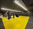 A spacious gym area with a yellow and blue padded mat, several hanging boxing bags, a few chairs, and a small cabinet with various items.