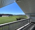A spacious balcony with white railings and a covered roof, overlooking a green field and distant mountains under a clear blue sky.