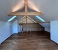 An empty attic space with wooden floors, white walls, exposed wooden beams, skylights, and a ladder leading to another level.