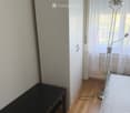 A small, furnished room with a wooden floor, a white wardrobe, a black table, and a floor lamp near the window with sheer curtains.