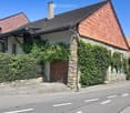 Two-story house with stone walls, red-tiled roof, and wooden front door, partially covered in ivy and surrounded by greenery.