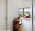 A modern, well-lit living space with a wooden floor, a small wooden table with a wicker basket and decorative items, a red sofa, and a view of a balcony area with outdoor furniture.