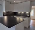 Modern kitchen with white cabinets, black countertop, and a kitchen island. The space features a sink with a faucet and an open doorway leading to another room.