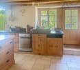 Bright kitchen with wooden cabinets, stainless steel appliances, and a large sink. Features a tiled floor, natural light from windows, and decorative elements like a flower vase and hanging herbs.