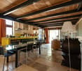 Open-plan living and dining area with exposed wooden beams, a glass dining table with chairs, a sofa, a floor lamp, and a kitchen area with a counter, refrigerator, and shelves.