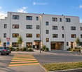 Modern two-story building with white facade, multiple windows, and a covered entrance. A car is parked nearby, and a pedestrian crossing is visible in front.