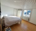 Bright bedroom with wooden flooring, slanted ceiling, large window offering a view, bed with white bedding, nightstand with books, and a wardrobe.