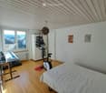 A multi-functional room with a bed, exercise bike, keyboard, and a cabinet. The room features wooden flooring, white walls, and large windows with a scenic view.