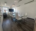 Modern conference room with wooden flooring, white walls, and large windows. Features a glass table surrounded by white chairs and a large screen mounted on the wall.