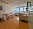 Modern office space with wooden flooring, large windows, a sofa, office chair, desk, and a screen.
