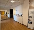 Reception area with a white reception desk, brochures, a chair, a small table, and a potted plant. The floor is wooden, and the ceiling has recessed lighting.