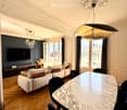 Modern living room with a white sofa, marble dining table, dark blue chairs, large windows with curtains, and a television on a wooden stand.