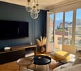 Modern living room with a grey sofa, coffee table, side table, television, and pendant lights. The room features large windows with a scenic mountain view and a balcony accessible through glass doors.