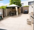 A modern outdoor patio with beige tiles, a covered seating area with a table and chairs, a grill, and a fire pit surrounded by stone walls.