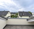 Roof terrace with grey tiled surface, white textured walls, and metal railings. The view shows neighboring houses with solar panels on their roofs and a cloudy sky.