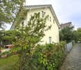 A well-maintained garden area with a variety of plants and trees, a small table and chairs, and a pathway leading to a two-story house with a light yellow facade and multiple windows.