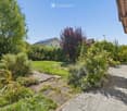 Well-maintained garden with a variety of plants, shrubs, and trees, featuring a paved pathway leading to a wooden bench near the house.