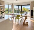 Bright, modern kitchen and dining area with light wood flooring, white cabinetry, and large windows overlooking a scenic outdoor space. Features a central dining table with colorful chairs, a high chair, and a kitchen island with a gas stove.