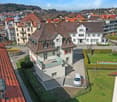 Aerial view of a residential neighborhood with multiple houses, red-tiled roofs, and a mix of architectural styles. A white car is parked in a driveway, and there is a small garden area with a swimming pool and potted plants.