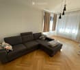 Living room with a grey sectional sofa, patterned pillows, wooden floor, large windows with curtains, radiator, and modern pendant lights.
