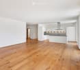 Open-plan unfurnished room with light wooden flooring, white walls, and a modern kitchen area featuring white cabinets, a stainless steel range hood, and a black countertop.