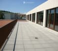 Modern rooftop terrace with light grey tiled flooring, metal railing, and large windows overlooking a residential area.