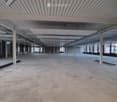 Empty industrial-style room with concrete floor, exposed white beams, and large windows allowing natural light.