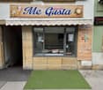 Facade of a gelateria named "Me Gusta" with a striped awning, glass display window, and menu board listing soft drinks and coffee options.
