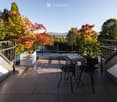 Balcony with a round table and four black metal chairs, surrounded by trees and a clear view of the street.