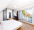 Bright bedroom with wooden flooring, large sliding glass doors leading to a balcony, a bed with white linens and red pillows, a mirrored wardrobe, a small table and chair, and a view of greenery and mountains.