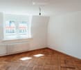 Empty room with wooden herringbone flooring, white walls, large window with white frame, radiator below window, and a single light fixture hanging from the ceiling.