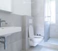 Modern bathroom with a white sink, chrome faucet, toilet, and glass-enclosed shower. Grey tiled walls and floor.