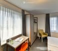 A modern, furnished room with a bed, a desk, a chair, and a wardrobe. The room features large windows with curtains, a grey carpeted floor, and a contemporary design.