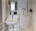 Modern bathroom with a white sink, mirror, bathtub, and showerhead. Includes a towel on the edge of the tub and a small shelf with toiletries.