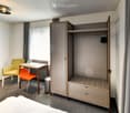 A modern, compact bedroom with a bed, a wardrobe, a small table with chairs, and a stool, all set in a cozy, well-lit space.