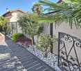 A well-maintained garden area with a paved walkway, decorative white pebbles, small trees, shrubs, and a wrought-iron railing leading to a house entrance.