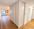 A clean, modern hallway with light wooden flooring, white walls, and a large sliding glass door leading to a balcony area with city views.