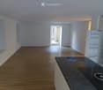 Empty room with wooden flooring, white walls, and a partially open door leading outside. A countertop with a small appliance and a few items is visible on the right side.
