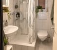 Modern bathroom with white tiles, a bathtub with a shower curtain, a sink with a mirror, a toilet, and decorative plants on the windowsill and shelf.