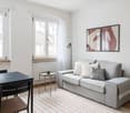 Bright and airy living room with large windows, grey sofa, black dining table, and abstract artwork on the walls. The room has a cozy and modern feel with a striped rug on the floor.