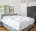 Spacious bedroom with a gray upholstered bed frame, white bedding, and a large black wardrobe. The room features framed artwork on the walls, a geometric pendant light, and a potted plant, creating a modern and minimalist aesthetic.