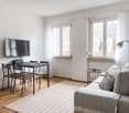 Spacious living room with large windows, hardwood floors, a dining table with chairs, a gray sofa, and a TV mounted on the wall. The room has a minimalist and modern design aesthetic.