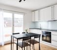Modern kitchen with white cabinets, black countertop, built-in oven, and dining table with black chairs. Large windows provide natural light and views of the surrounding buildings.