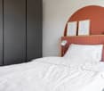 minimalist bedroom with white bedding, dark grey wardrobe, pink headboard, wall lamps, framed art, and potted plants